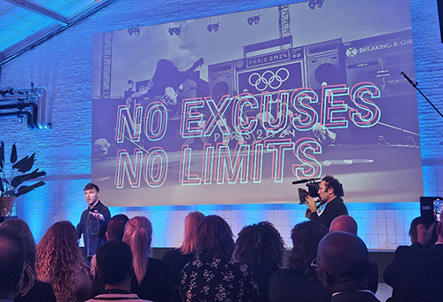 Redo staat op een podium. Een cameraman filmt hem. Op een scherm achter hem staat in grote letters No Excuses No Limits